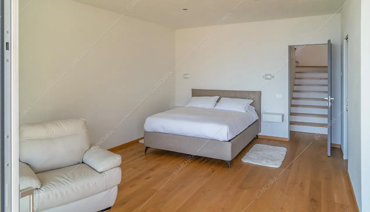 Camera da letto matrimoniale con poltrona bianca, pavimento in parquet, pareti bianche e vista sulle scale