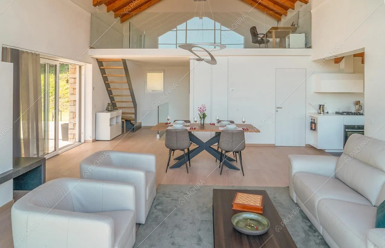 Salotto con divano e poltrone bianche, studio sul soppalco con balaustra in vetro e vista sulla cucina a muro