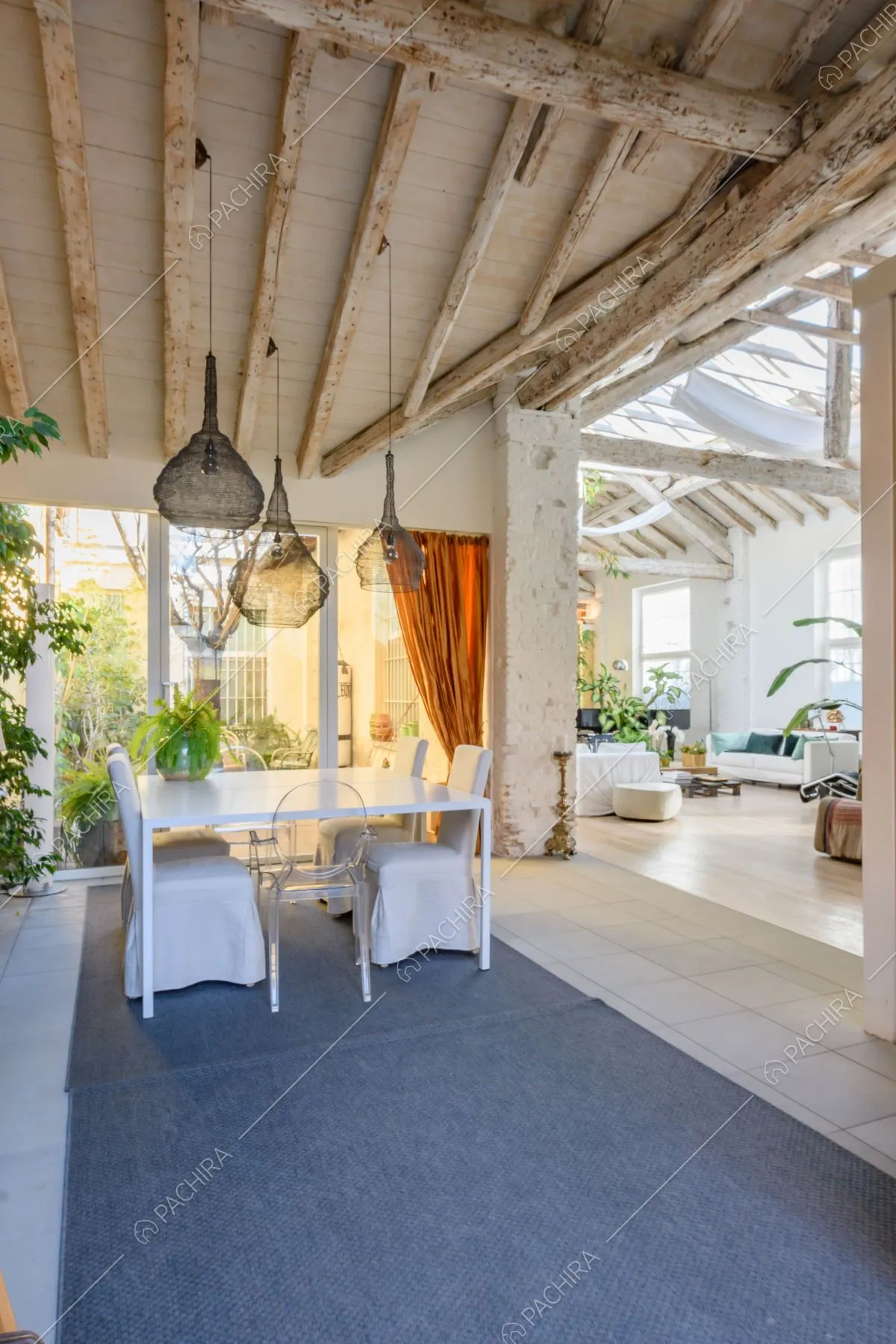 Tavolo da pranzo e sedie bianche nella veranda con porte finestre e travi in legno grezzo a vista sul soffitto