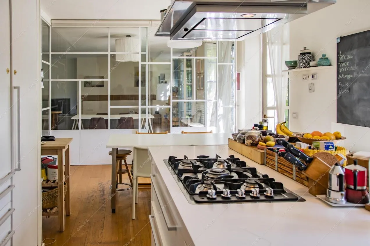 Cucina con fornelli a gas, vetrate industriali dagli infissi bianchi e vista sul salotto