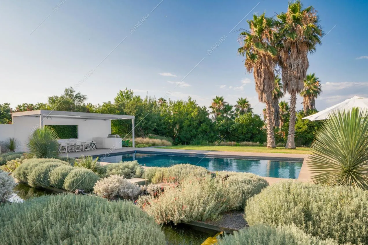 Dai cespugli del giardino vista sulla piscina e sulla veranda con tavolo da pranzo