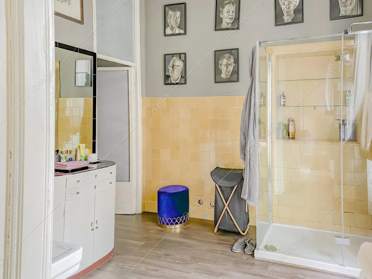 Pouf cilindrico in velluto blu, doccia con mensole e ritratti fotografici appesi alla parete