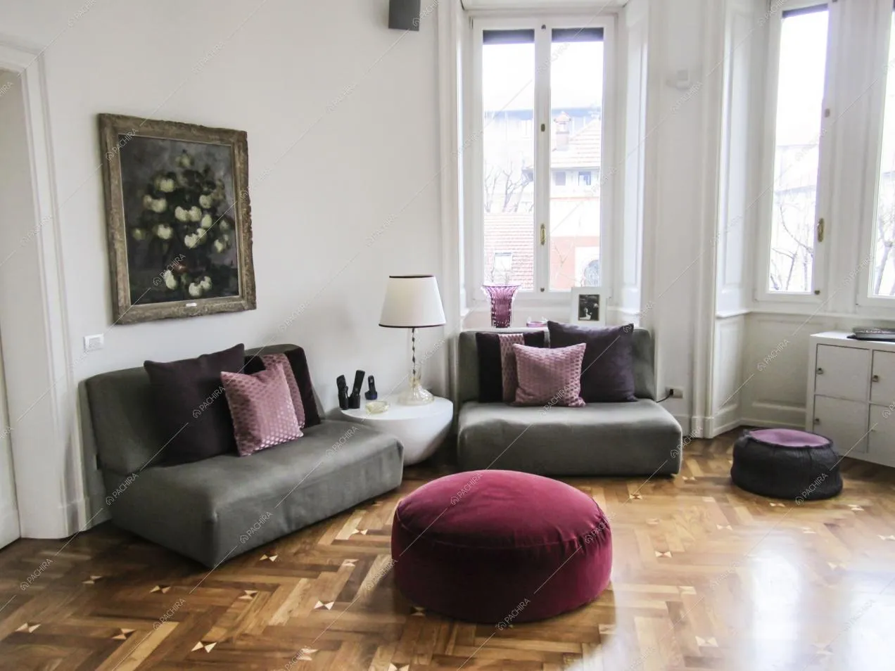 Luminoso soggiorno dalle pareti bianche con pavimento in parquet a spina di pesce, divani e pouf