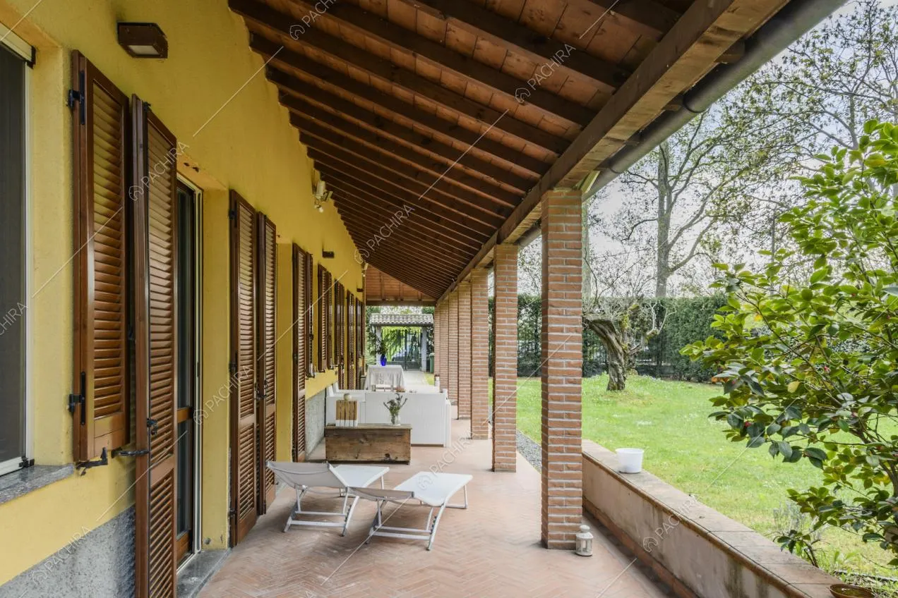 Lungo portico con travi a vista, persiane marroni e giardino con piante