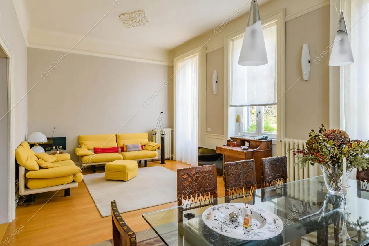 Sala con tavolo da pranzo, pareti grigie, boiserie sul soffitto, grandi vetrate e divani gialli