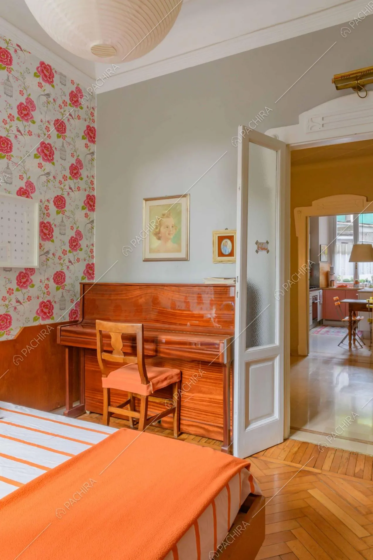 Camera da letto con carta da parati a fiori, pianoforte a muro e scorcio sulla cucina
