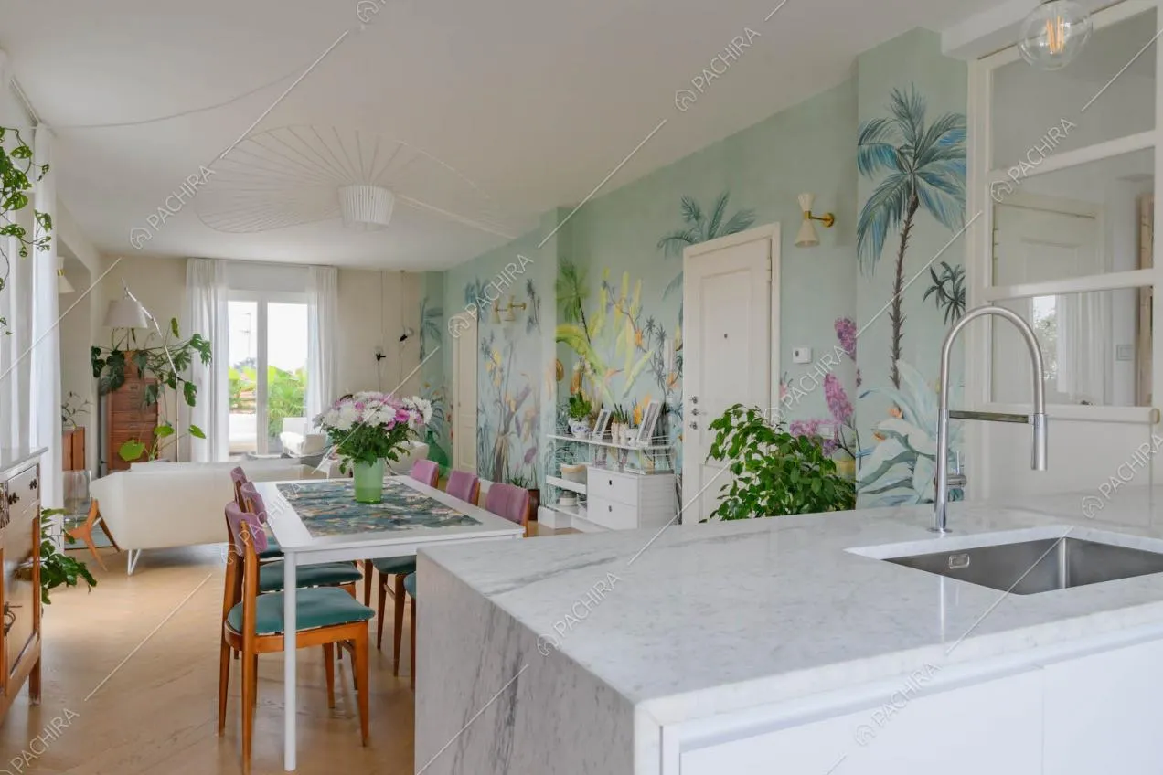Dalla penisola della cucina con piano da lavoro in marmo vista sul salotto con carta da parati a fantasia tropicale