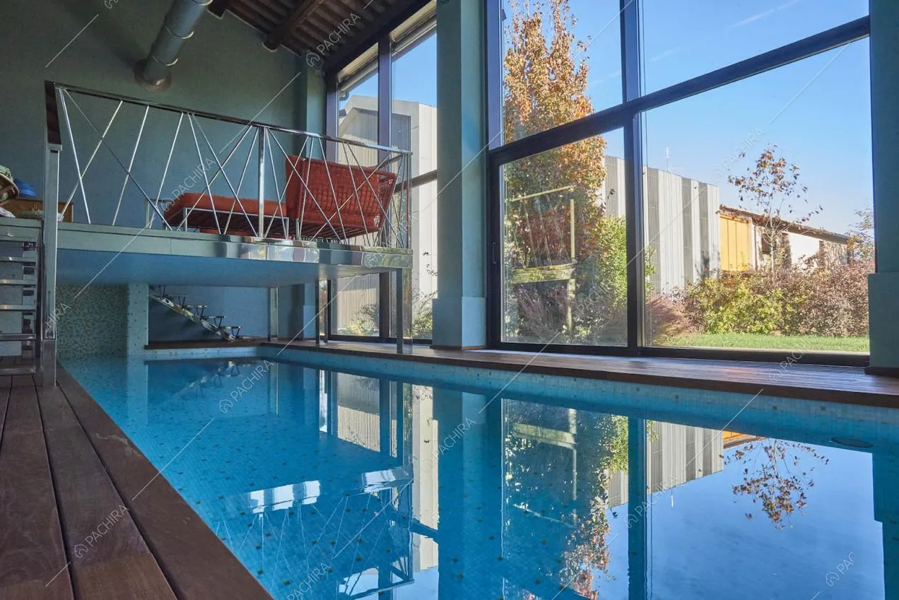 Piscina interna nella sala con soppalco, grandi vetrate e soffitto in travi in legno a vista