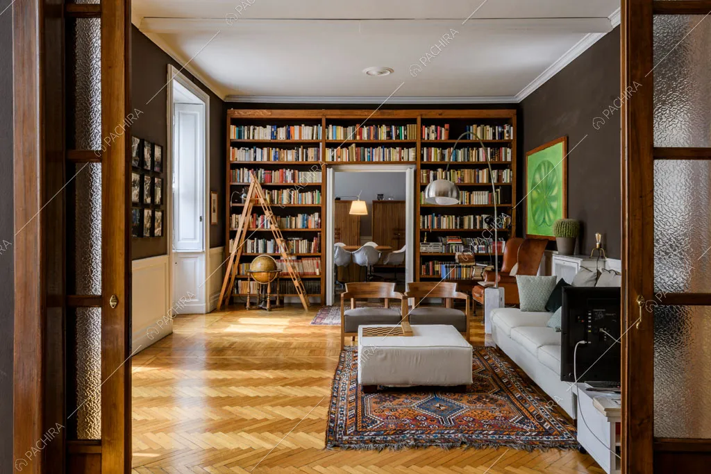 Dalla porta a vetri antica vista sulla sala con divano bianco, pouf e pareti marroni con boiserie