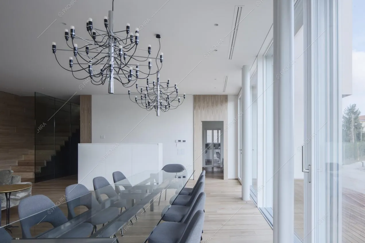 Lungo tavolo da pranzo con piano in vetro, illuminato dalle porte-finestre del salotto, e vista sulle scale in legno