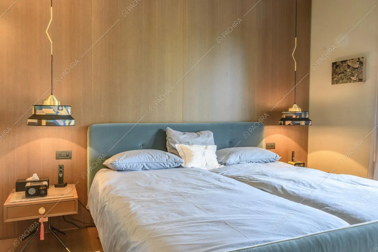 Letto matrimoniale con spalliera in velluto, comodini, lampade appese al soffitto e parete in legno