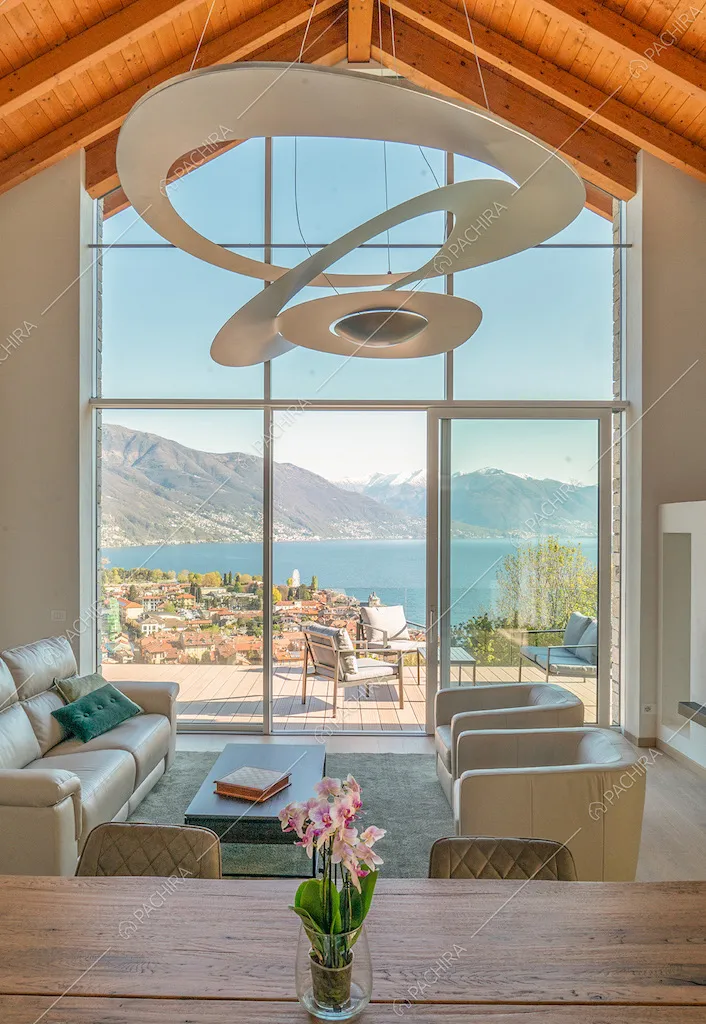 Soggiorno illuminato da un'ampia vetrata con tavolo da pranzo in legno, poltrone, divano e lampadario di design