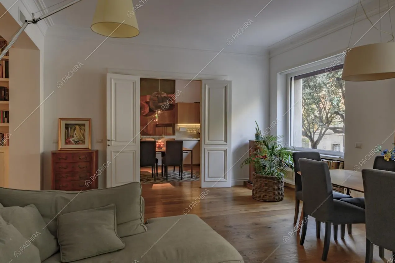 Salotto dalle pareti bianche con boiserie, pavimento in parquet e vista sulla cucina
