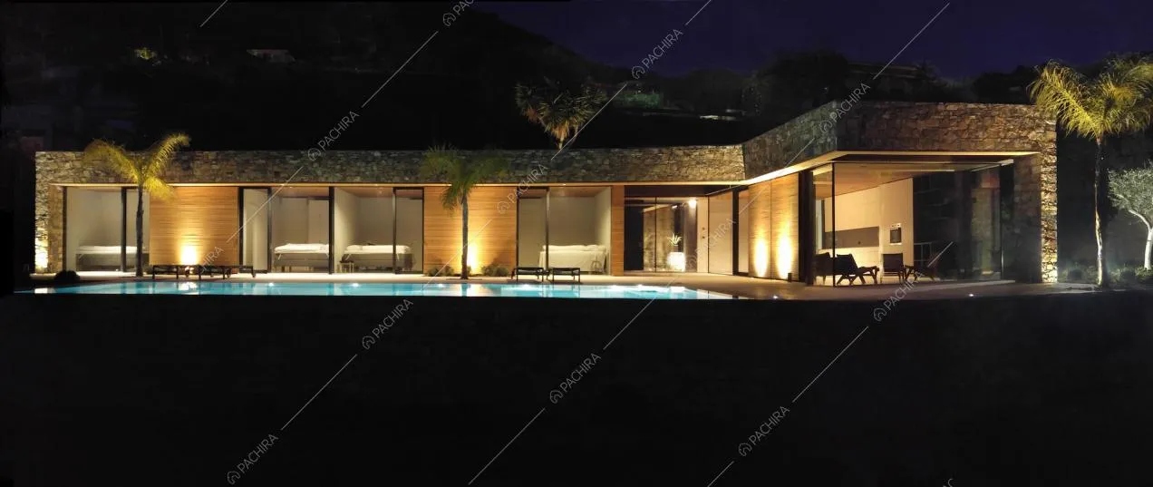 Di notte vista sulla piscina a sfioro illuminata e sugli esterni della villa, in cui si alternano vetrate a pareti in legno