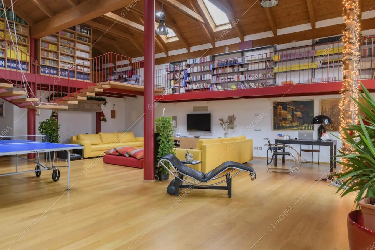 Salone con divani gialli, soppalco con libreria e soffitto con lucernari e travi in legno a vista
