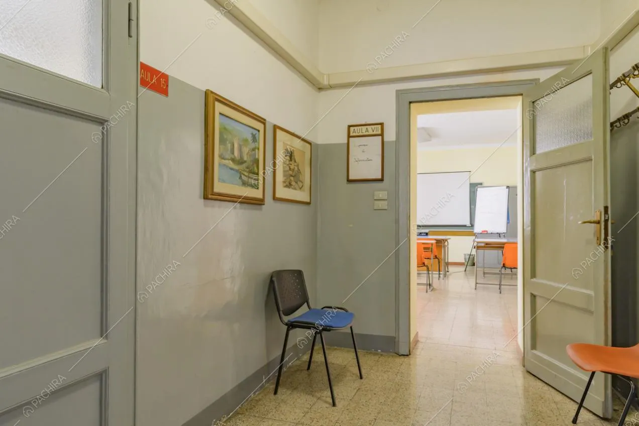 Dal corridoio, con attaccapanni al muro, vista su un'aula dalle pareti gialle e pavimento in piastrelle